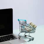 Mini shopping cart with rolled cash next to a laptop, representing online shopping.